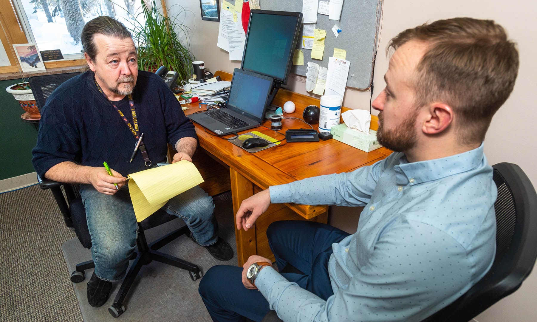 A man with a notepad talks to a nicely dressed young man in an office.