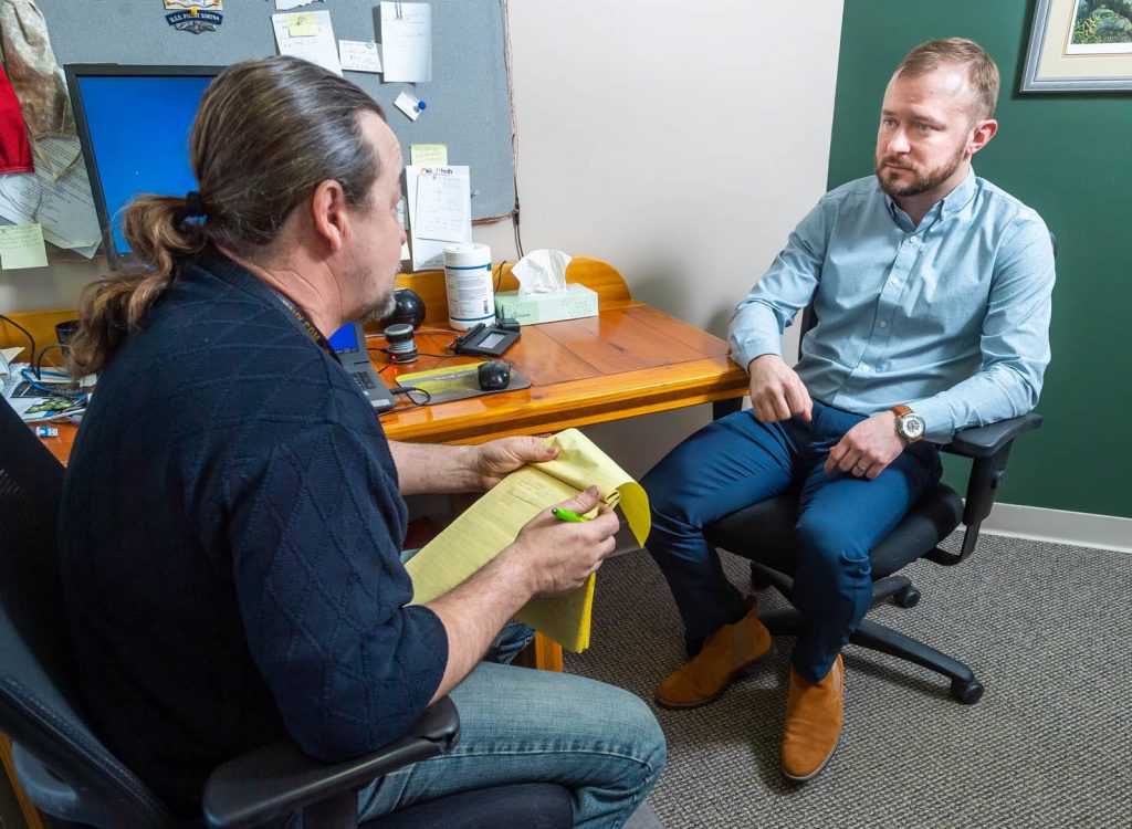 A man with a notepad talks to a nicely dressed young man in an office.