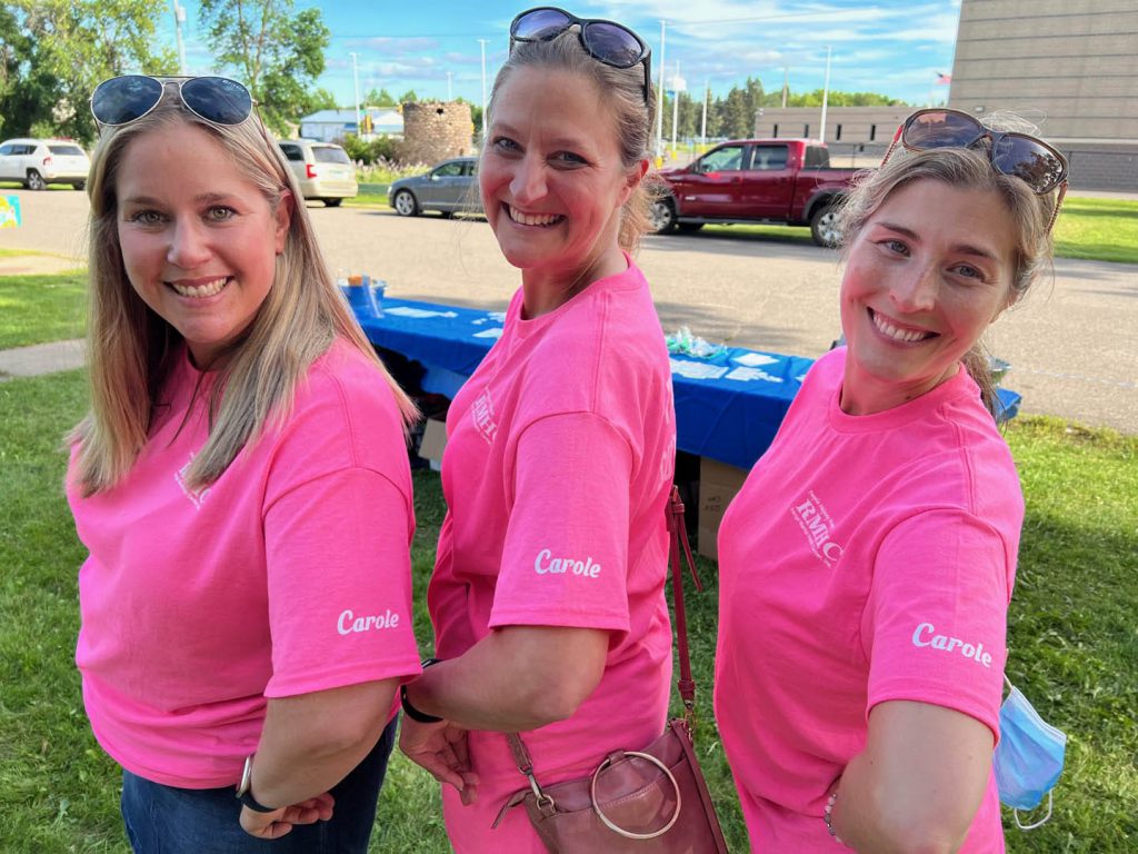 Three women in pink t-shirts smile at the camera at an outdoor event.