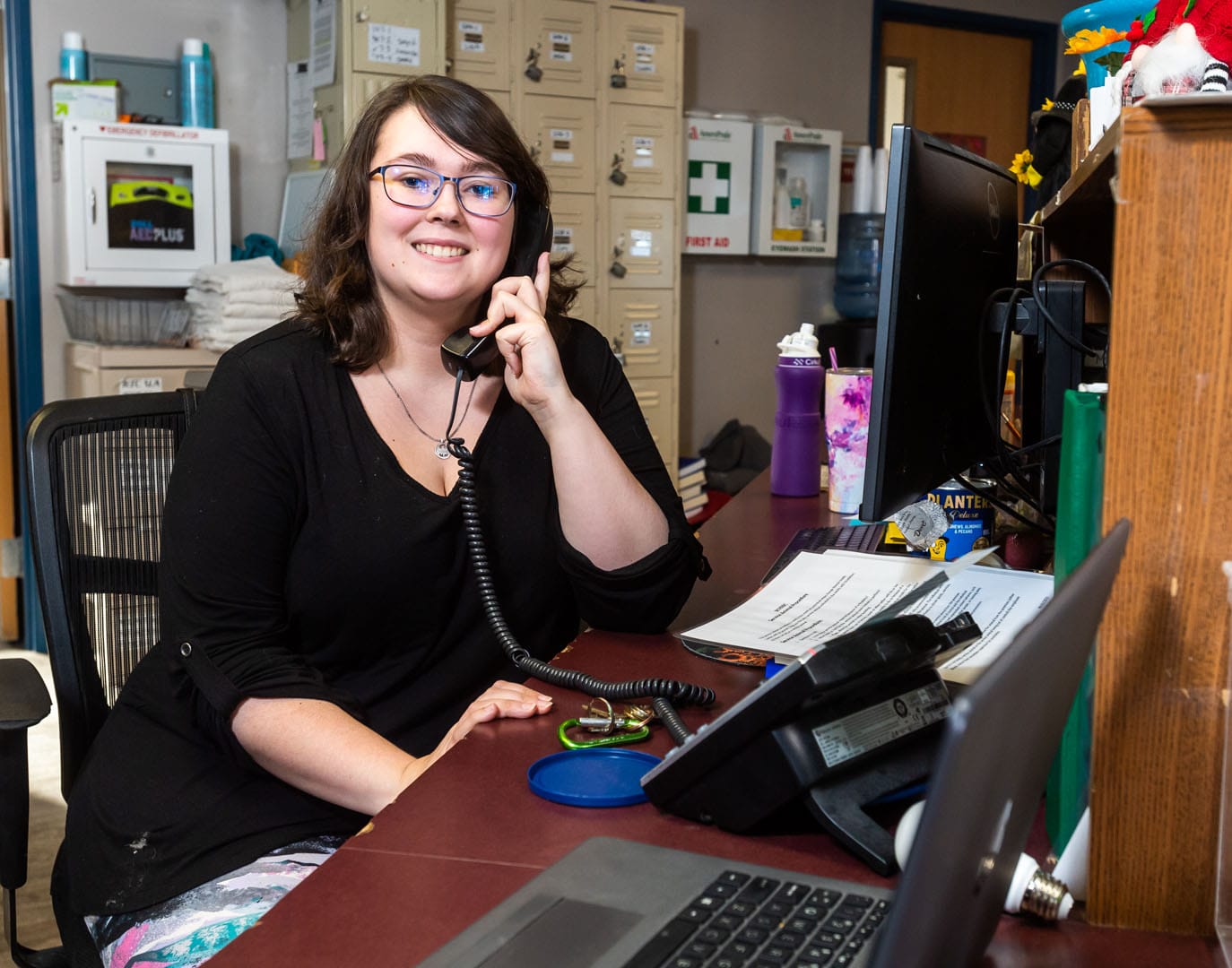 A smiling young woman sits at a desk and answers a phone.