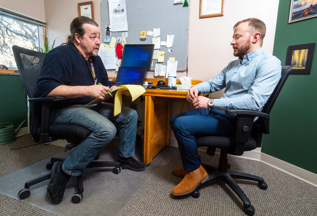 A man with a notepad talks to a nicely dressed young man in an office.