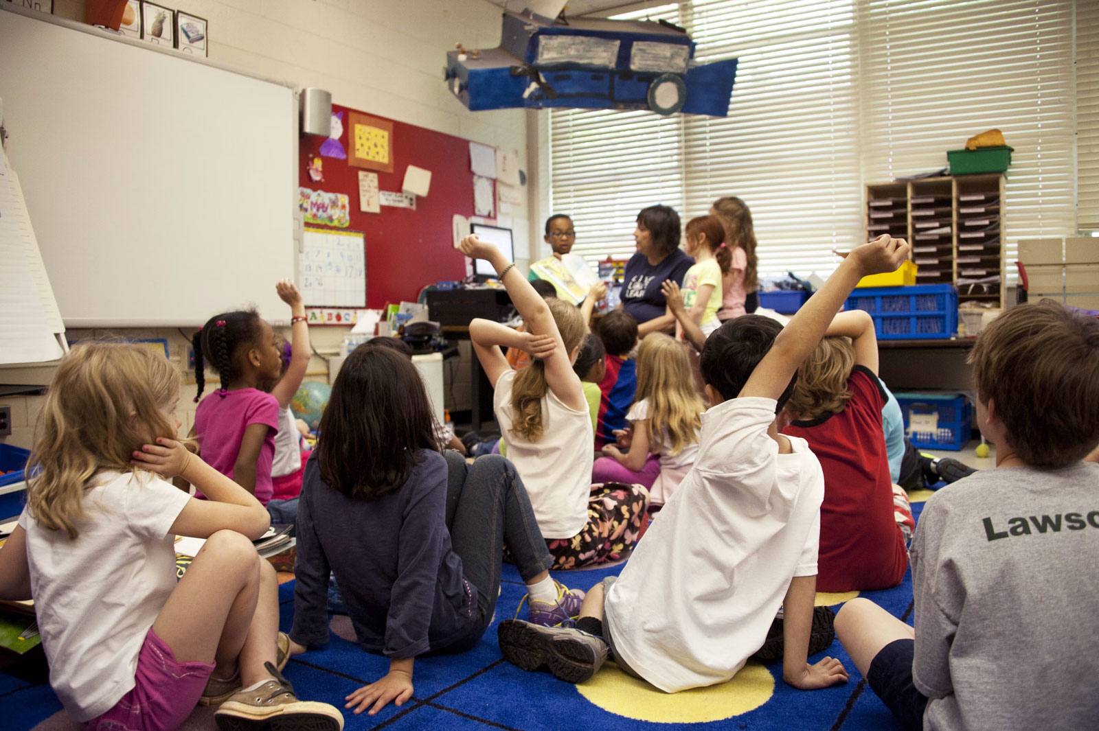 Young kids in an elementary classroom raise their hand while a teacher reads to them.